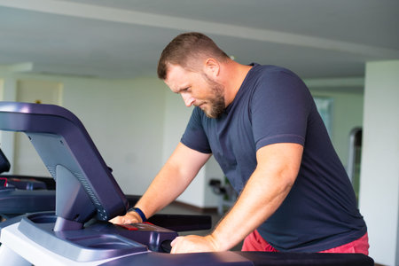 upset tired full male runs on a treadmill in a gym. concept of weight loss and sport. side view