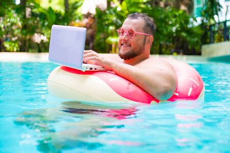 Fat funny man in pink inflatable circle in pink glasses works on a laptop in a swimming pool.の写真素材