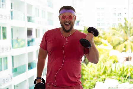 Funny fat male in pink glasses and in a pink T-shirt engaged with dumbbells in the gym portraying a girl.の写真素材