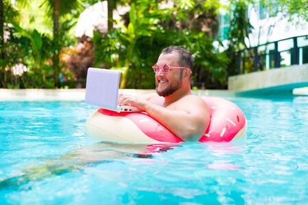 smiling Fat funny man in pink inflatable circle in pink glasses works on a laptop in a swimming pool.の写真素材