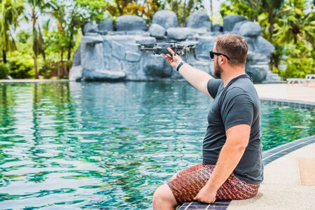 Bearded man launches drone for flight, with which you can take photos and video filming, near the swimming pool and palm trees, side viewの写真素材
