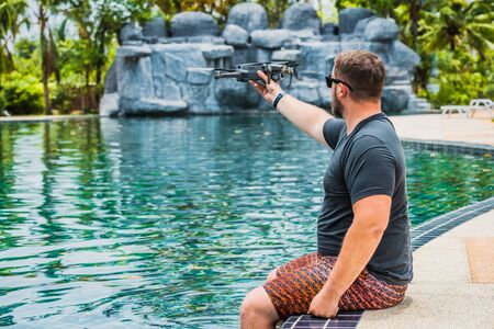 Bearded man launches drone for flight, with which you can take photos and video filming, near the swimming pool and palm trees, side viewの写真素材