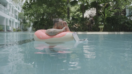 Fat funny man in pink inflatable circle in pink glasses works on a laptop in a swimming pool. The man is surprised and drops the laptop into the water in the poolの写真素材