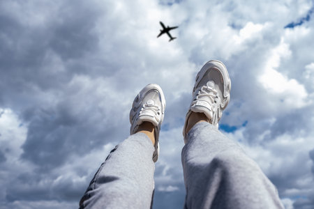 Legs in white sneakers against the background of a cloudy sky and a flying airplane. Freedom conceptの写真素材