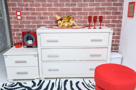 A white chest of drawers on a background of a brick orange wall, next to a red armchair and a...