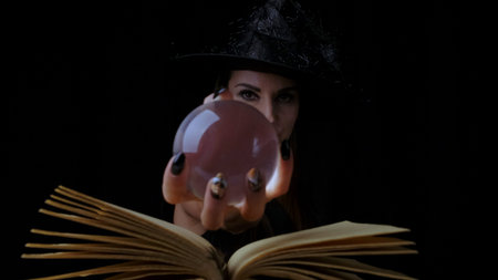 female in a black witch costume and hat, holds in her hands a transparent magic ball over a book and...