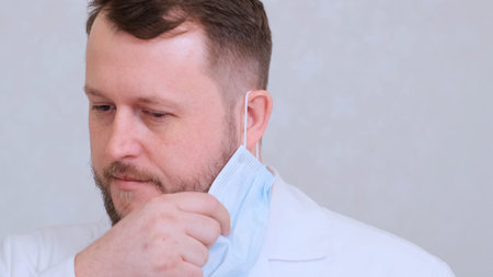 bearded male doctor takes off his protective mask with a sad look. This is the end. Hygiene concept....