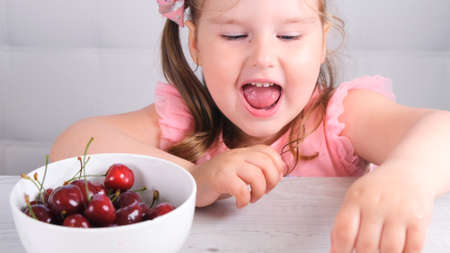 Beautiful cute little girl sitting at a light wooden table with a plate of cherries and eating a berry. Healthy eatingの写真素材