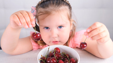 little girl sitting at a light wooden table with a plate of cherries and eating a berry. Healthy eatingの写真素材