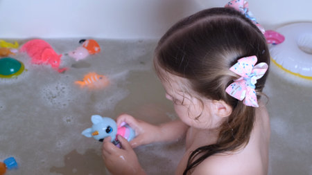 Little girl 3 years old with two ponytails on her head, bathes and washes in the water in the bathroom at home. Baby body care concept, hygieneの写真素材