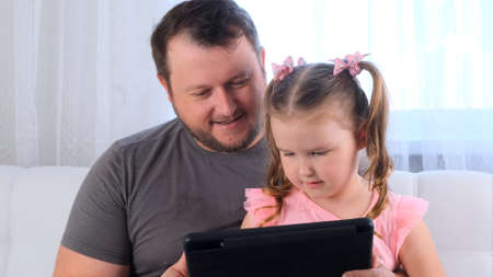 interested little girl 3 years old and her father study together on a tablet and watch a lesson online at home. Dad helps his daughter with her studies. Online school distance learning conceptの写真素材