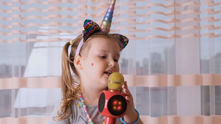 funny cute little girl 4-5 years old, singing into a karaoke microphone, with a unicorn headband, child singing karaoke music, have fun at an event future musician loud voice solo.の写真素材