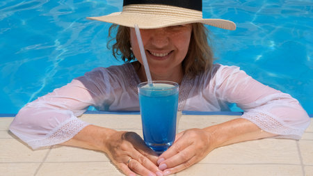 Smiling senior woman relaxing near the blue outdoor swimming pool with a blue cocktail. People are...