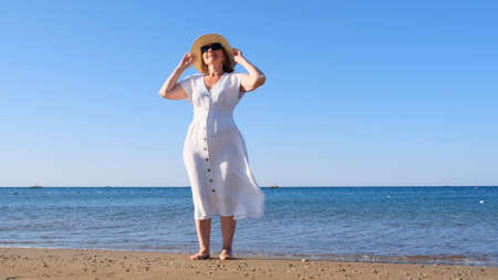 mature woman in sunglasses in a straw hat and white dress walks along the blue sea coast on a sunny summer day, enjoying freedom and relaxation. The concept of a senior citizen everyday life.の写真素材