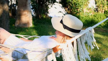 relaxed senior woman in straw hat smiling happy relaxing on a hammock enjoying the fresh air on the terrace around the palm trees near the sea. Senior citizen lifestyle concept.の写真素材