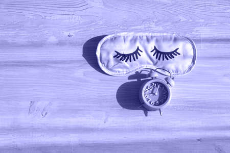 alarm clock and sleep mask on a wooden white table in the morning sunlight. The concept of starting a new day, good morning or ending the day, late evening. Day planning. Very Peri toned image. copy space.の写真素材