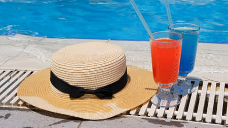 Two colorful cocktail glasses pink and blue, sun hat on the edge of the swimming pool, close up. vacation and relaxation concept in spa hotel.の写真素材