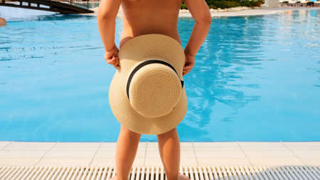 sweet little girl 4 years old with a straw hat dances and looks at the blue sea or pool, back view, close-up. Childhood, relationship, summer vacation concept. selective focus on the hatの写真素材