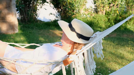 relaxed senior woman in straw hat smiling happy relaxing on a hammock enjoying the fresh air on the terrace around the palm trees near the sea. Senior citizen lifestyle concept.の写真素材