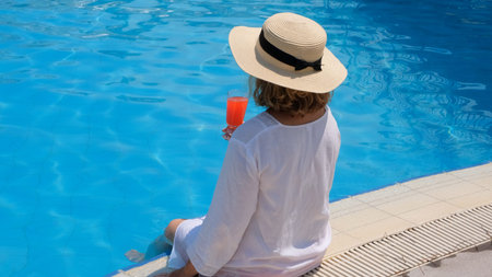 fashionable blonde woman in straw hat relaxes and drinks orange cocktail near the swimming pool on a...