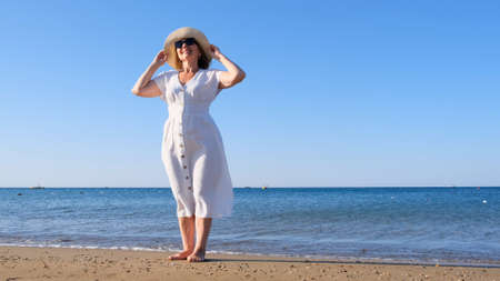 woman in a straw hat and white dress walks along the seashore and admires the blue sea on a sunny summer day, enjoying freedom and relaxation. The concept of a senior citizen everyday lifeの写真素材