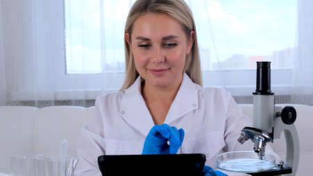 Woman scientist research chemist in a medical suit and protective gloves with a tablet computer and a microscope conducts research in the laboratoryの写真素材