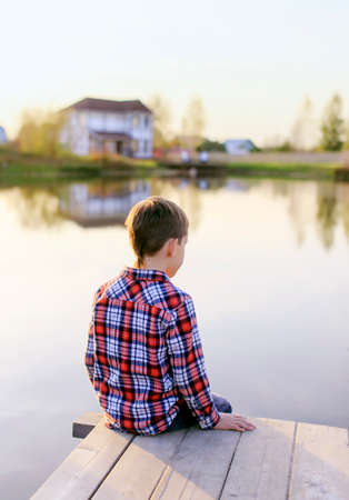 boy looking at the water pond. thoughtful boy in plaid shirt looking at smooth water of the pond. On the opposite bank is visible two-storey houseの写真素材