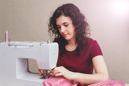 Happy attractive young woman seamstress sitting and sews on sewing machine in studioの写真素材