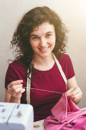 Happy attractive young woman seamstress sitting and sews on sewing machine in studioの写真素材