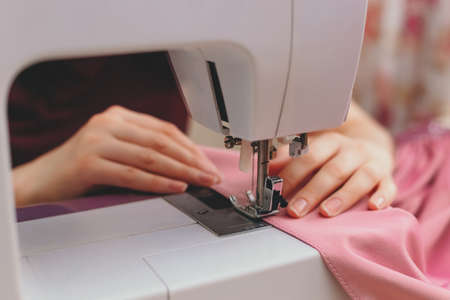 Happy attractive young woman seamstress sitting and sews on sewing machine in studioの写真素材