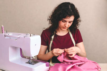 Happy attractive young woman seamstress sitting and sews on sewing machine in studioの写真素材