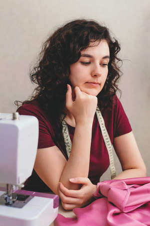 Happy attractive young woman seamstress sitting and sews on sewing machine in studioの写真素材