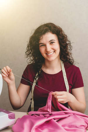 Happy attractive young woman seamstress sitting and sews on sewing machine on light background. Concept of sewing, handmade goods. Advertising of studio. Sunlightの写真素材