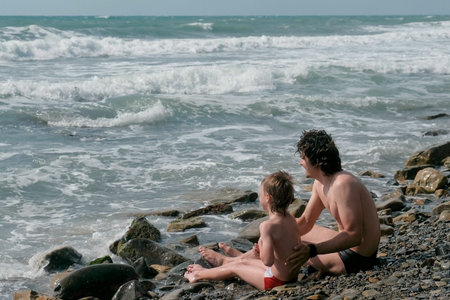 Dad and son is sitting in sea waves in storm on stone beach. Back view