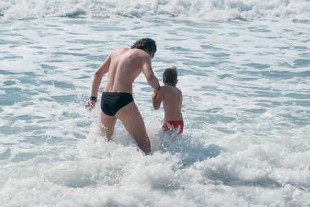 Dad and son go to swim in sea waves in storm on stone beach. Back viewの写真素材