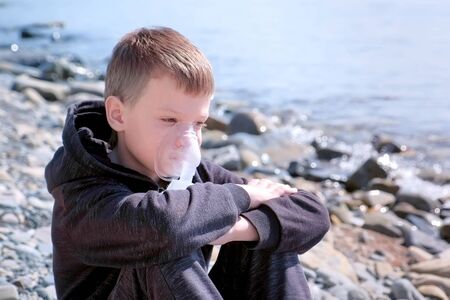 Portrait sick 8y child boy uses nebulizer sits on seaside. Inhaling inhaler mask. Fibrosis cystic copd and treatment. Asthma pulmonary respiratory breath problem cure. Painkiller sedative gas oxygen.の写真素材