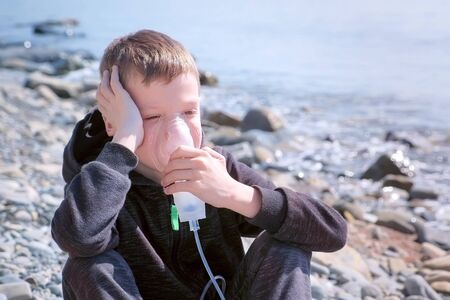 Portrait sick 8y child boy uses nebulizer sits on seaside. Inhaling inhaler mask. Fibrosis cystic copd and treatment. Asthma pulmonary respiratory breath problem cure. Painkiller sedative gas oxygen.の写真素材