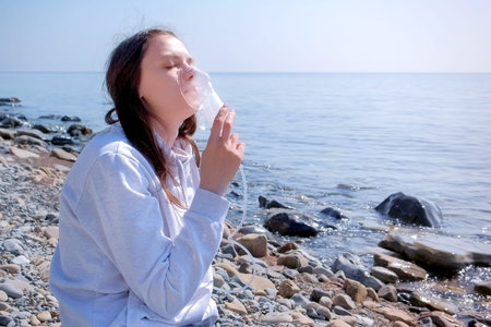Young sick woman uses nebulizer on seaside. Inhaling inhaler mask. Fibrosis cystic copd and treatment inhaler. Asthma pulmonary respiratory breath problem and cure. Painkiller sedative gas oxygen.の写真素材
