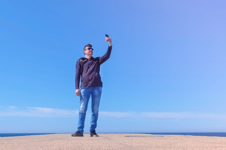 Young man tourist travel blogger shooting video on smartphone at sea pier, low angle view. Guy...