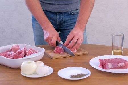 Man cutting fresh fillet meat pork on pieces to cook shashlik, hands closeup. He cutting on wooden board kitchen table. Prepare cook food for grilling on grill. Spices, butter and onions on table.の写真素材
