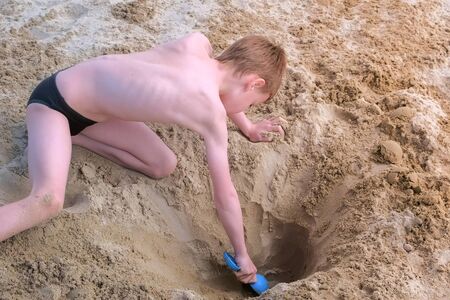 Child boy playing on sand beach digging shovel deep hole and throwing sand on sea ocean coast. Outdoor summer activities at vacation. Boy is sunbathing in swimming trunks.の写真素材