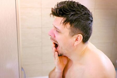Portrait of sleepy young man yawning and looking at mirror in bathroom in morning, side view. Trying to wake up. Lack of sleep, insomnia and stressful lifestyle. Hangover. Midlife crisis, depression.の写真素材