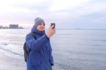 Man blogger talking video chat on smartphone walking at sea and showing beach in winter near coastal city. Beautiful nature at sunset. Tourist travel journey trip with backpack.の写真素材