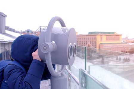 Unrecognizable child boy looking through binoculars on observation deck on roof of building in city in winter. Portrait of teen traveler in warm jacket and hat. Travel, journey and tourism concept.の写真素材