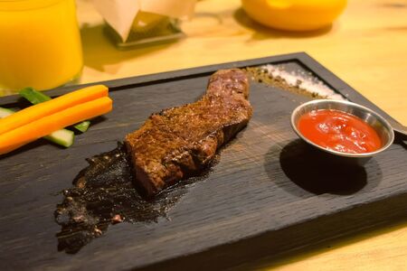 Piece of medium-roasted beef served on a black wooden Board with vegetables and tomato sauce in a restaurant. Sea buckthorn herbal tea at table. Tasty dinner.の写真素材