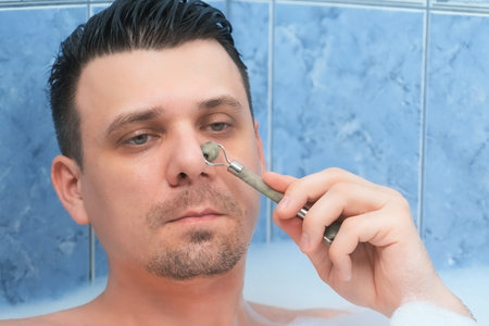 Portrait of healthy fresh handsome bearded man model massaging skin with face jade roller massager performing morning routine. He is relaxing lying in bath and doing face massage himself.の写真素材