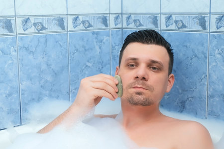 Portrait of healthy fresh handsome bearded man model massaging skin with face jade gouache scraper massager performing morning routine. He is relaxing lying in bath and doing face massage himself.の写真素材