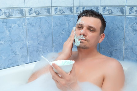 Young man is applying professional alginate mask lying in bathroom with foam at home. Spa beauty treatment. Man wellness, skincare and self love concept. Relaxing and enjoying his time.の写真素材