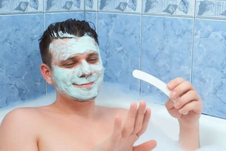 Young man is filing his nails sitting in bathroom with mask on face. Spa beauty treatment at home. Man wellness, skincare and self love concept. Relaxing and enjoying his time.の写真素材