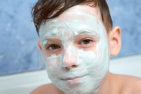 Portrait of child teen boy with alginate mask on face looking at camera and smiling. Spa beauty treatment at home in bathroom. Teen problem skin, wellness, skincare and self love concept.の写真素材